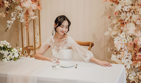 身穿蕾絲白紗的新娘坐在桃園扶日婚禮空間的長桌前拍攝婚紗,白橘色系花藝與金色燭台點綴,氛圍浪漫溫馨,適合婚宴使用