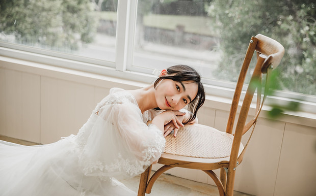 新娘身穿蕾絲白紗優雅倚坐在窗邊木椅上,陽光灑落室內,婚禮場地的高挑長窗營造輕奢溫柔氣質,適合婚紗拍攝使用