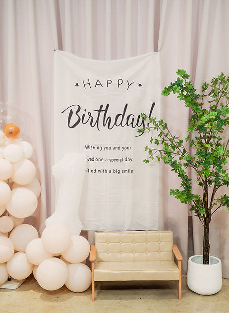 溫馨生日派對背景佈置，粉膚色氣球柱與透明氣球點綴，白色 Happy Birthday 布旗、綠植與米色沙發營造輕柔浪漫氛圍