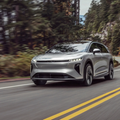 Silver Lucid Gravity SUV driving on a winding forest road. Motion blur emphasizes speed. Yellow lines on asphalt, trees in background. Dynamic mood.