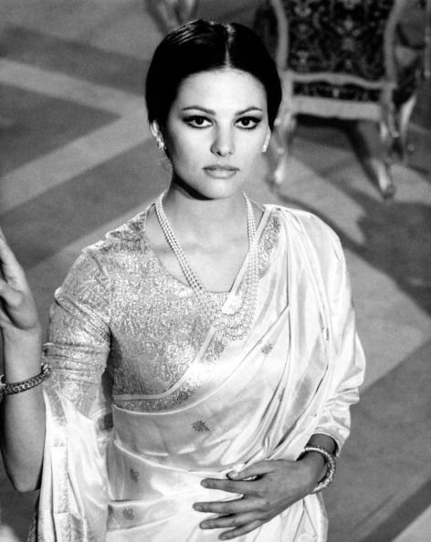 Claudia Cardinale, in an elegant saree with intricate embroidery and pearl necklaces, standing in a lavish room, conveying grace and poise.