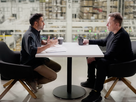 Elon Musk and Nikhil Kamath sit across from each other at a table in a warehouse, discussing with coffee mugs in hand. An American flag hangs in the background.