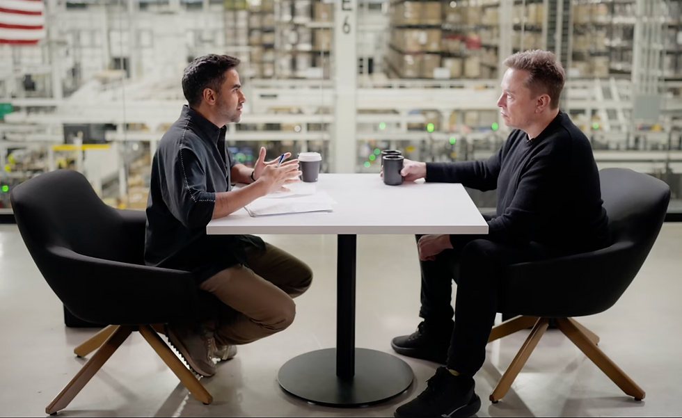 Elon Musk and Nikhil Kamath sit across from each other at a table in a warehouse, discussing with coffee mugs in hand. An American flag hangs in the background.