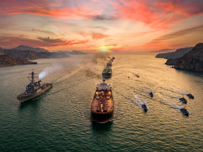 Ships sail through a narrow strait at sunset, flanked by escort vessels. Dramatic orange and pink sky contrasts with the calm green water.