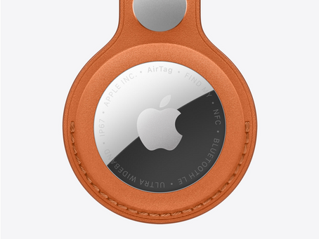 A silver Apple AirTag in a sleek, brown leather keychain. Visible text: "Apple Inc., AirTag, Find My, NFC, Bluetooth LE."