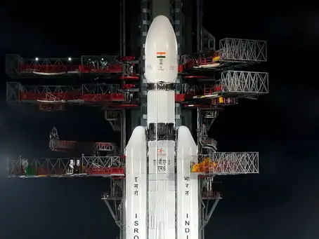 Rocket on launch pad at night, labeled ISRO and INDIA. Text above reads "LVM3 - M6 / BlueBird Block-2 Mission." Lights illuminate scene.