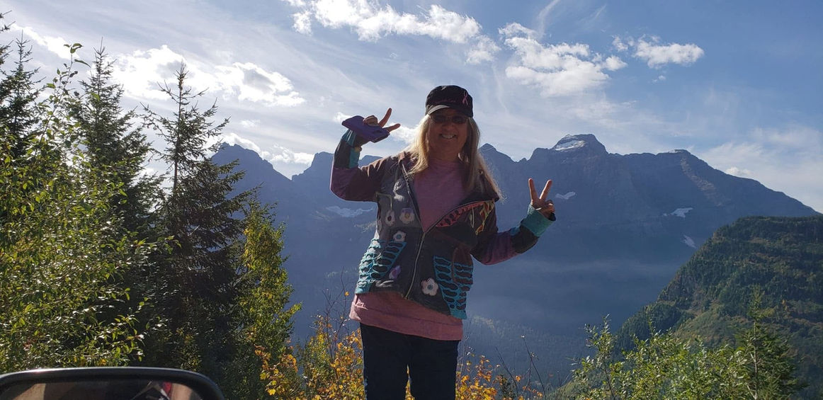 a blonde female standing infront of a mountain smiling holding up two "peace" hand gestures.