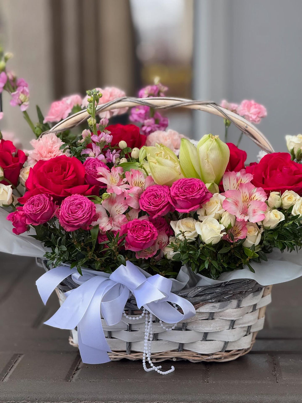 Basket of mixed pink and red roses, carnations, alstroemerias, and tulips, decorated with a white satin bow.