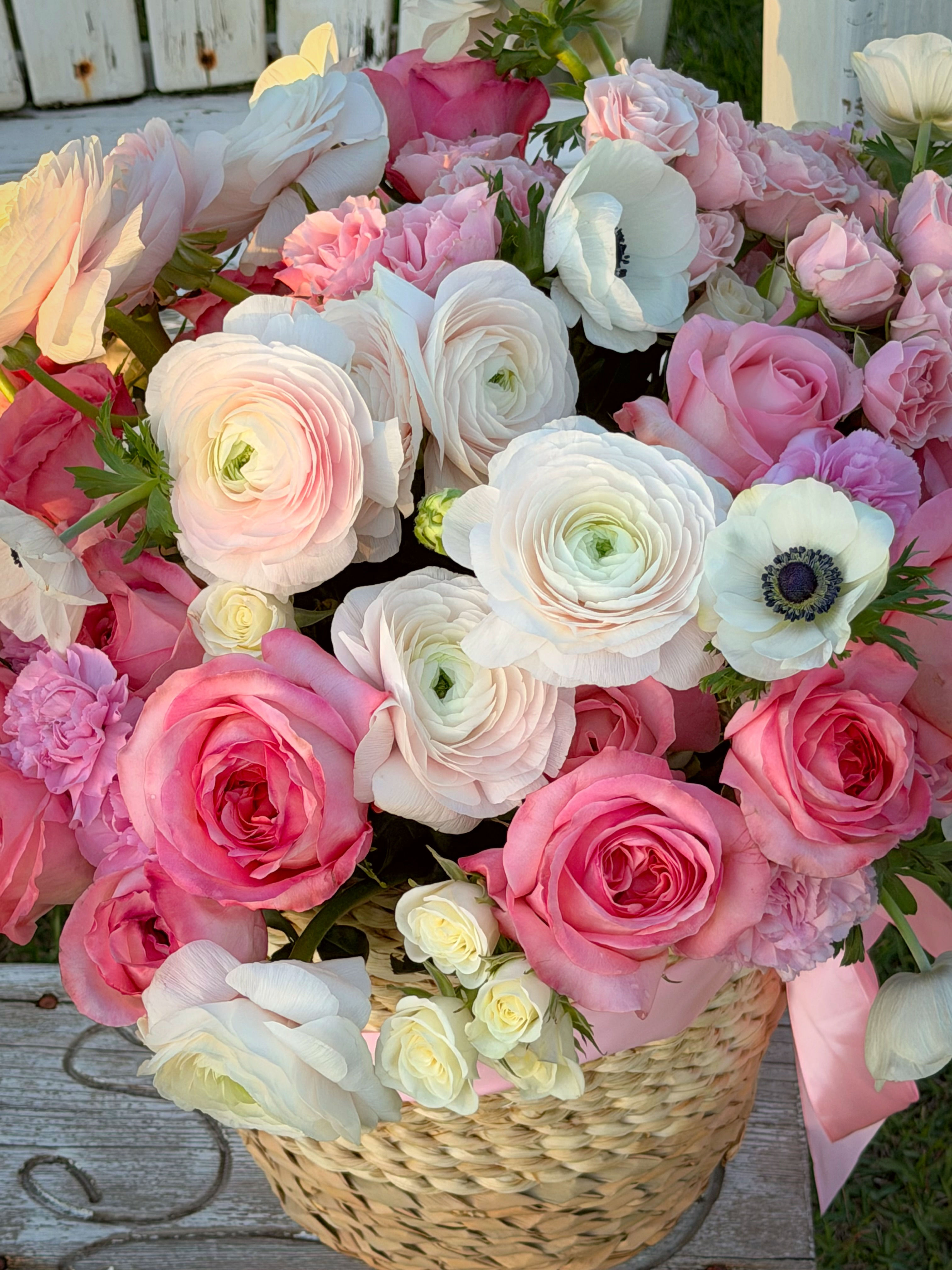 A romantic basket arrangement featuring pink roses, white ranunculus, and delicate accents in a natural woven basket. Soft, a