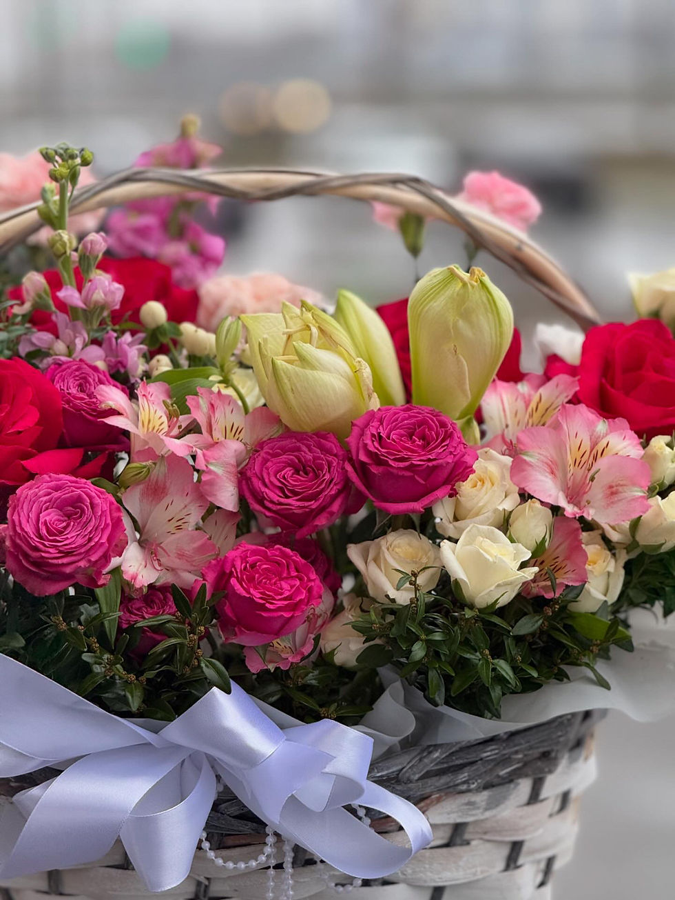Basket of mixed pink and red roses, carnations, alstroemerias, and tulips, decorated with a white satin bow.