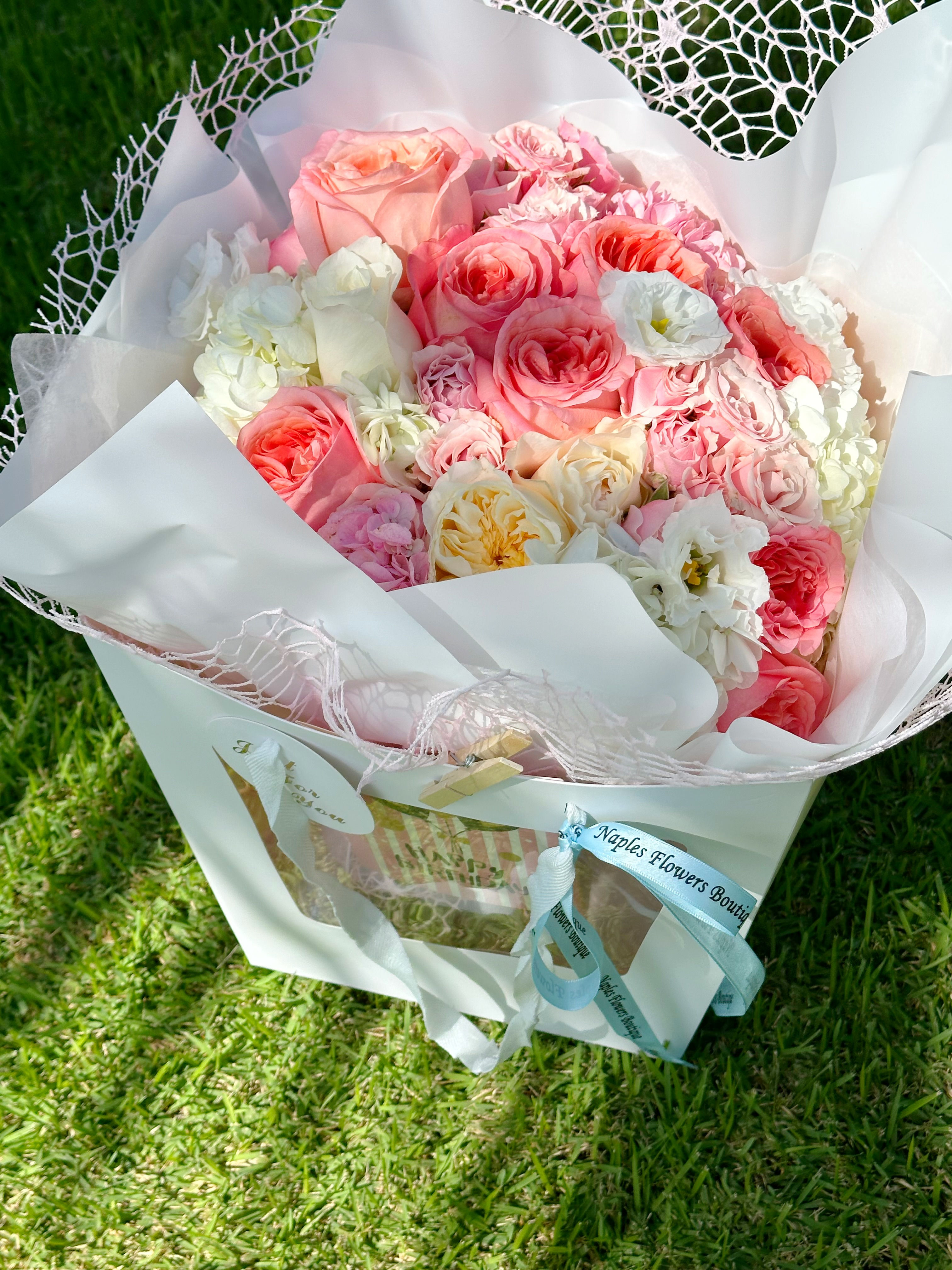 Soft pastel bouquet with pink roses, lisianthus, and hydrangeas in white gift bag wrap.