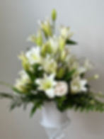 Arrangement of white lilies and roses in a white container, surrounded by lush greenery.