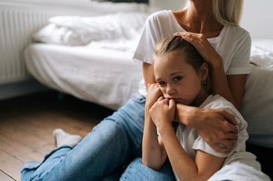 Child sitting on bed looking worried while parent sits nearby offering comfort.