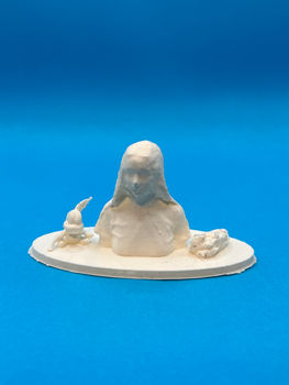 A 3D-print of myself placed on an oval base, shown against a bright blue background. The figure is simplified and abstracted, with minimal facial detail and a softly textured surface, accompanied by small sculptural forms on either side.