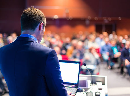 a man standing infornt of a laptop and microphone speaking t a crowd of people in the background