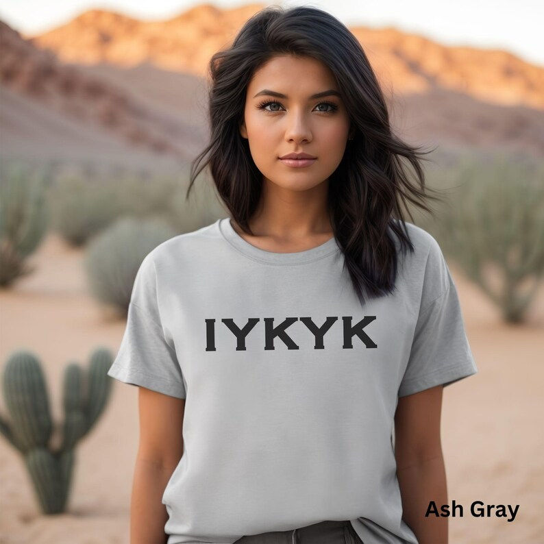 A woman stands in a desert landscape wearing an ash gray T-shirt featuring the bold "IYKYK" logo, capturing the essence of the brand in a serene natural setting.