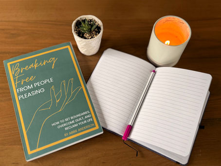 Book titled Breaking Free next to an open notebook with a pen, a small potted plant, and a lit candle on a wooden table.