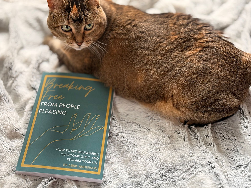 Tabby cat lying on a gray blanket next to a book titled "Breaking Free From People Pleasing" by Abbie Anderson.