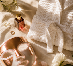 flat lay of luxury Mother's Day gifts including a white robe, perfume bottle, and rose gold ring on a cream surface