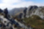 Hiker with backpack on rocky trail overlooking jagged mountains under cloudy sky. Rugged terrain, earthy tones evoke adventure and solitude.