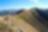 Mountain ridge under a clear blue sky, with dry grass and rocky terrain. Distant mountain ranges create a serene and expansive view.