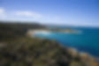 Lush green hills meet blue ocean under a clear sky. Waves crash on rocky coastline, dotted with sandy beaches. Calm and scenic.