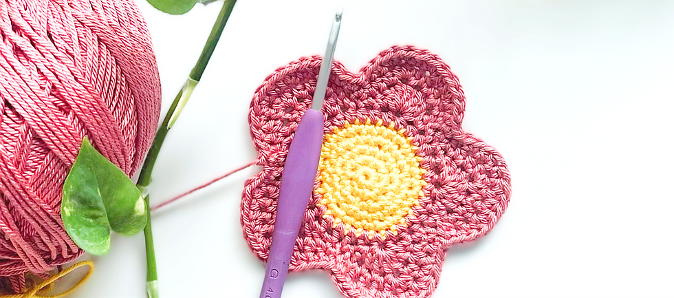 Flor tejida con hilaza rosa y el ovillo rosa al lado adornado con una pequeña planta. El circulo del centro amarillo y el ganchillo con