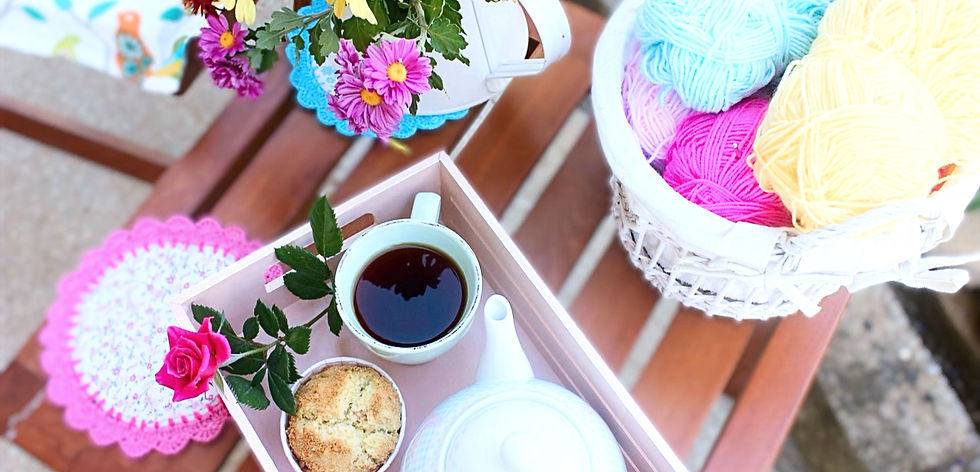 Una mesa de madera con una caja de madera que tiene dentro una tasa de cafe, un pankake y una tetera, al lado una canasta con hilazas, al lado un florero con flores y
