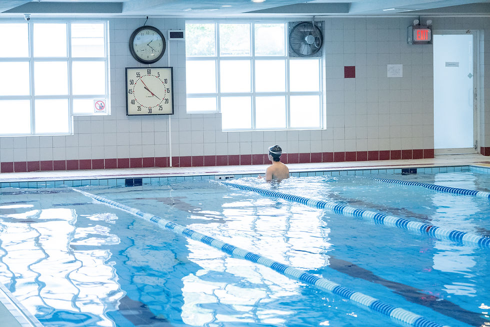 Swimmer resting at the edge of an indoor lap pool with lane dividers, wall clocks, bright windows, and an EXIT sign.