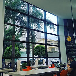 A imagem mostra o interior de um café ou espaço de trabalho com grandes janelas de vidro que permitem a visão de palmeiras e edifícios do lado de fora. Dentro do local, há mesas brancas com cadeiras de cores variadas, incluindo laranja e preto. À direita, uma pessoa está sentada em uma das mesas, usando um laptop. No canto superior direito da imagem, há uma parede com um design gráfico que inclui um desenho de uma lâmpada pendurada e texto, mas o texto não é legível na imagem. A cena é iluminada pela luz natural que entra pelas janelas, criando um ambiente claro e arejado.