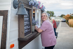 photo of an older woman in a purple shirt standing in front of a coffee trailer that is decorated with flowers