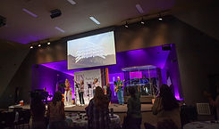 photo of 3 women and a male base player singing and playing a song on the church stage. The crowd is in the foreground with hands raised