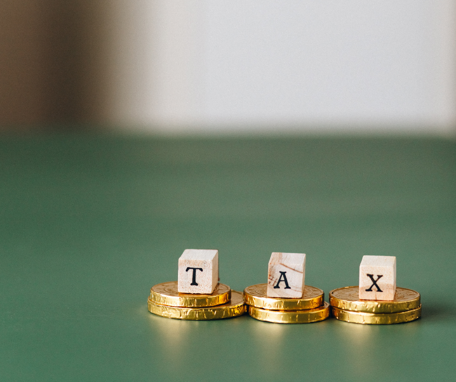 Blocks spelling "TAX" sit atop stacks of gold coins on a green surface, against a blurred background.