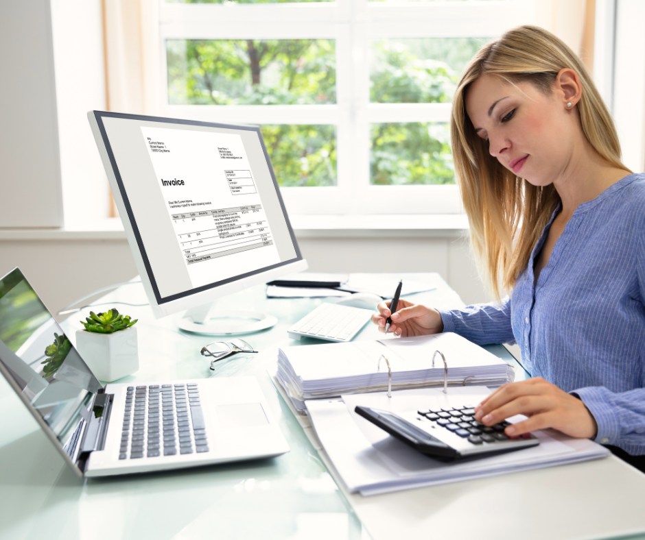Woman in blue shirt calculates with a calculator and writes beside an invoice on her desktop computer in a bright office.