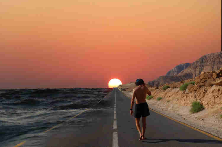 A man walking along an empty road towards the setting sun with the desert to the right and the sea to the left