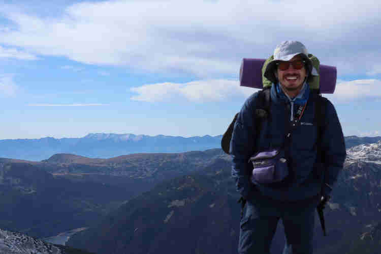 Oleh Nikita Smelyanskiy standing on top of a mountain smiling with a backpack, sleeping mat, and white hat