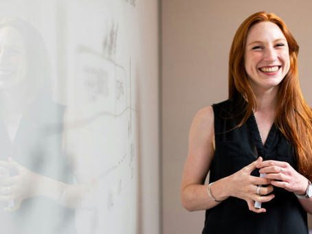 A woman with red hair smiles brightly while standing near a whiteboard, suggesting she is giving a confident presentation or discussion in a professional setting.