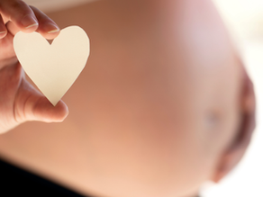 Close-up of a hand holding a paper heart near a pregnant belly. Soft light, calm mood. Logo text: Louisville Prenatal Massage.