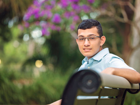 A guy sitting on a park bench with a tree with purple flowers in the background.
