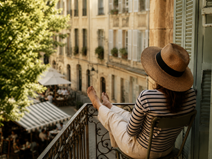 Woman relaxing on a sunlit balcony overlooking a quiet European street, capturing the peaceful, unhurried feeling of a French Sunday.