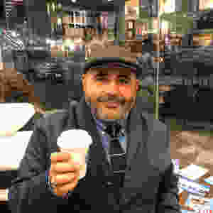 Ted wears a dark gray newsboy cap while dressed in a dark gray button front jacket, wearing a blue button-down shirt with a green horizontal striped tie and holding a small cup of coffee in a shop in front of large glass windows.