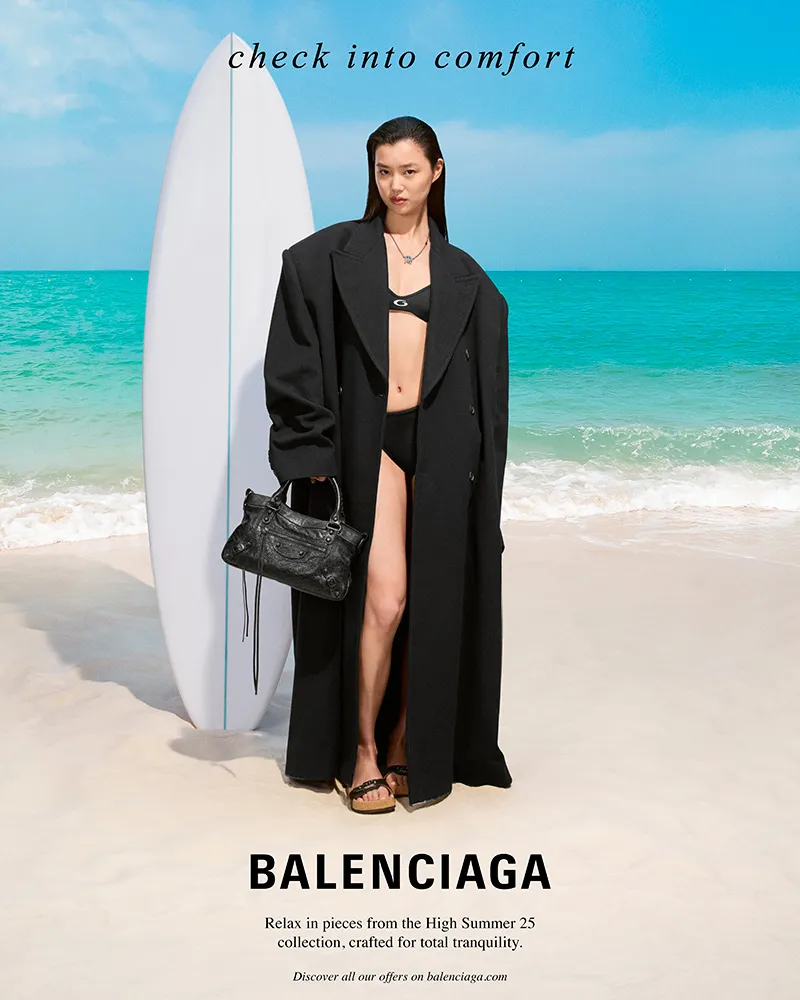 Balenciaga - High Summer 2025 campaign - a woman at a beach with crystal clear waters and white sand stoof in front of an upright white surf board wearing a full length black duster coat and bikini 