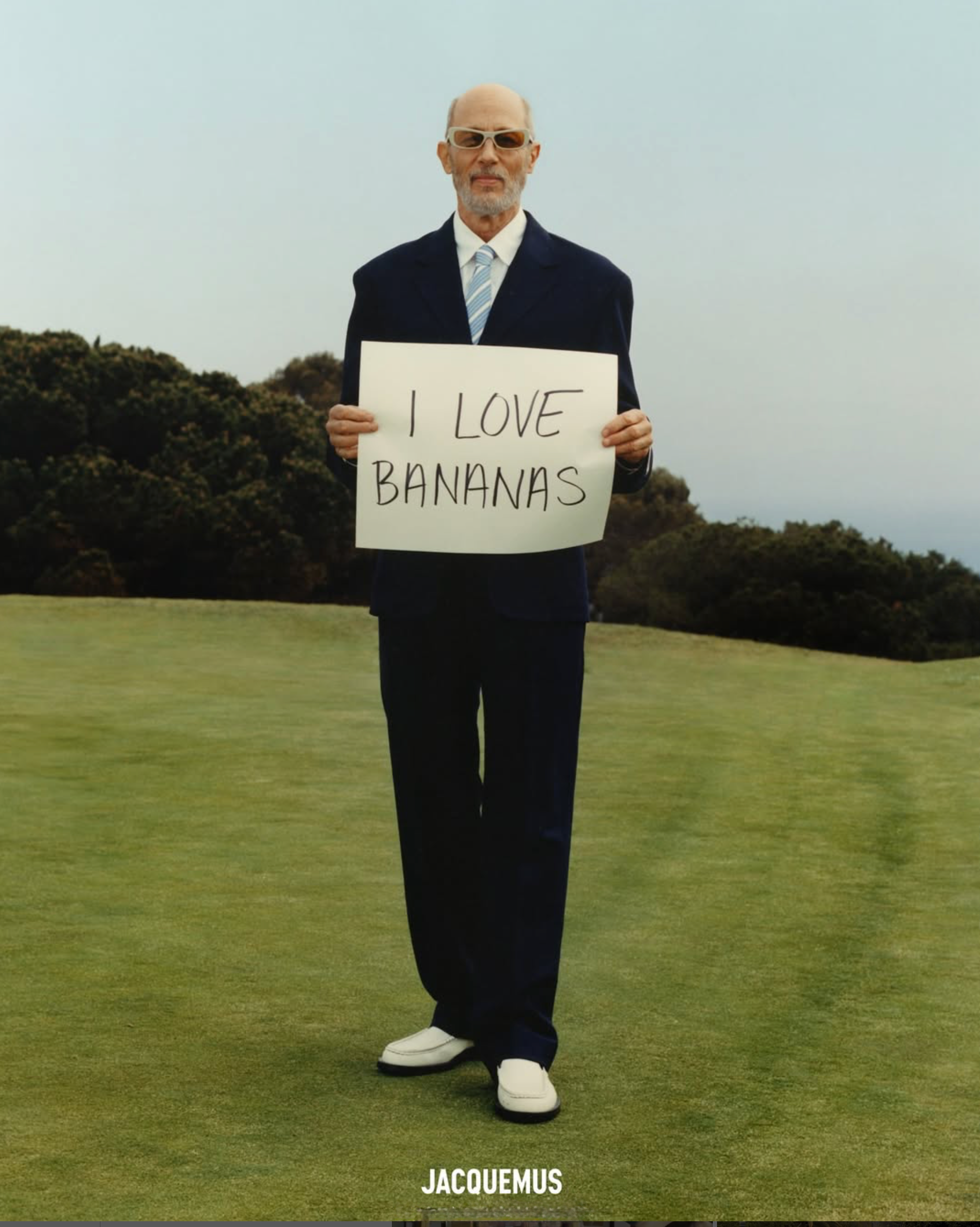 Jon Gries in a suit on a golf course holding a sign saying 'I LOVE BANANAS'