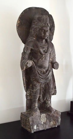 Hight 64 cm
In this full figure image sculptured in a dark chist, the Buddisattva or the Budda in the future, stands barefooted on a pedestal with his head surounded with a large halo. The missing right hand was probably raised in abbaya mudra position, and the left hand holding his charcteristic water pot.
He is wearing a turban withseries of retaining bandsemanating from a large barel bead. A thick , flowing hear goes down tho his shoulders and his moustasche has a double twist over a indistictly shaped mouth.
A long necklace separated by a large barrel bead in the middle. An amulet thpasses across the body with attached cylindrical amulets. A double armband on the right arm with an amulett picturing a small Buddha. His garmet is folded over his left shoulder and lifted over the right underarm. The pidestal is broken in the front, but has lotus rosettes on the sides that went around the statue.