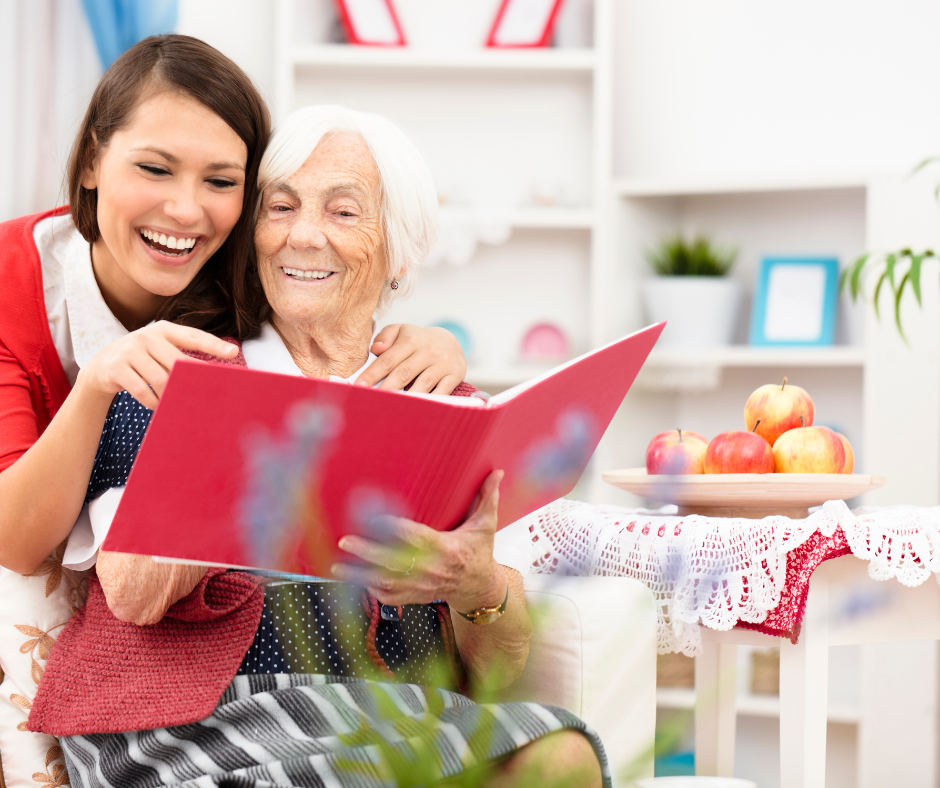 An image of a younger women with long straight brown hair. She is reading over the shoulder of a grey hair older women and they are reading from a book. The older woman is sitting in a comfortable chair smiling enjoying her time with the younger woman. They are both smiling and laughing. There is a small white side table with a bowl of fresh red apples. Bookshelf with picture frames are in the back of the room.