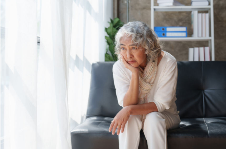Elderly woman in white sits pensively on a sofa by a window. Sunlight filters in, highlighting her thoughtful expression. Shelves behind.