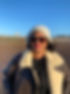 a photo of Maria, the coach at casulo. She is a brown skinned woman wearing bright pink lipstick. she is also wearing a white/grey beanie, sunglasses and a brown sheepskin aviator jacket. she is standing on the beach which you can just about see in the background, including the bright blue winter sky.