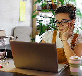 Young female gardener in glasses using laptop, communicates on internet with customer in h