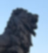 Bronze lion statue with a fierce expression, mouth open, against a clear blue sky. Its detailed mane is highlighted in the sunlight.