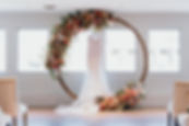 wedding dress hanging on a circular floral arch inside a venue. The arch is decorated with a variety of colorful flowers
